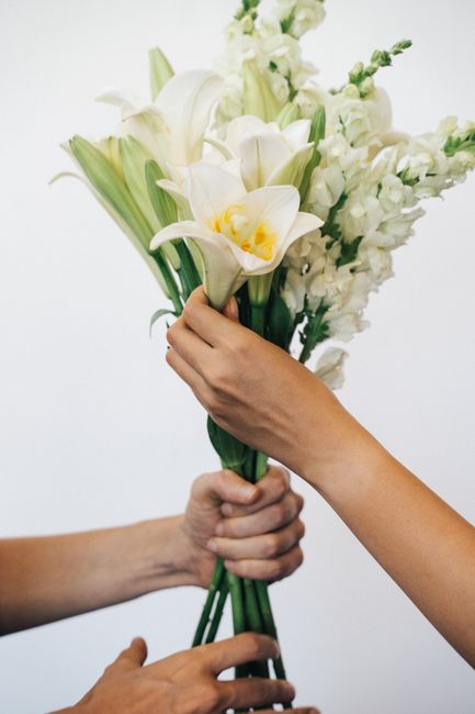 A close-up of two hands exchanging a bouquet of fresh white flowers, including elegant white lilies with yellow accents and delicate snapdragons, arranged in a natural, loose style. The flowers are he