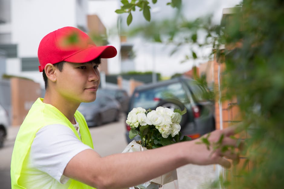 A young male florist from floristtemple.co.uk delivering a floral gift in a suburban residential area. He wears a red cap and a bright yellow vest over a white T-shirt, holding a bouquet of fresh whit