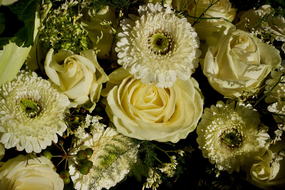 A floral arrangement featuring soft white roses, large cream-colored gerbera daisies with dark centers, and smaller white filler flowers, all arranged in a natural, lush bouquet. The flowers are set a