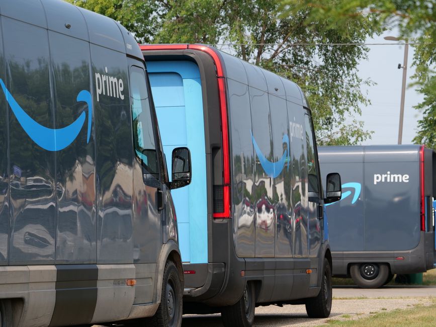 Image showing multiple Amazon Prime delivery vans parked outdoors with grey exteriors and prominent blue Amazon smile logos on each side. The vans are positioned on a street or parking area, with some