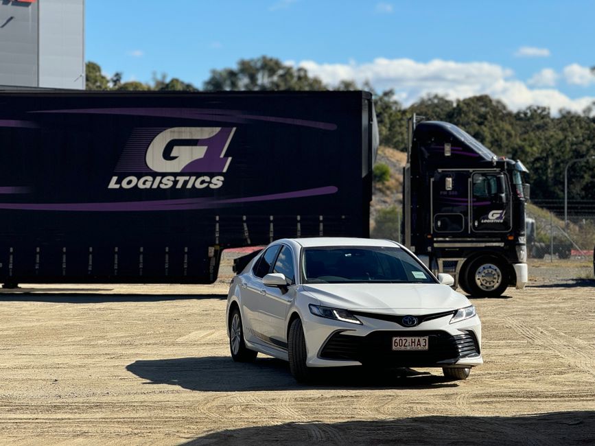 A white Toyota sedan is parked on a dirt surface in front of a large black G Logistics truck with purple accents, alongside a smaller black semi-truck cab, under a bright blue sky with scattered cloud